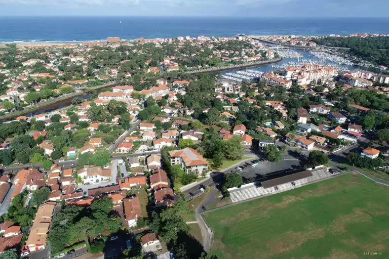 Cette image 3D est une vue aérienne d'une ville côtière ensoleillée. Au premier plan, on peut voir des zones résidentielles avec de nombreuses maisons aux toits de tuiles oranges, entourées d'une végétation dense. Au centre de l'image, un port de plaisance est visible, rempli de bateaux. Il est relié à l'océan par un canal. L'océan, de couleur bleue et calme, s'étend à l'horizon. Une plage de sable s'étend le long de la côte, et on peut voir des bâtiments côtiers derrière elle.