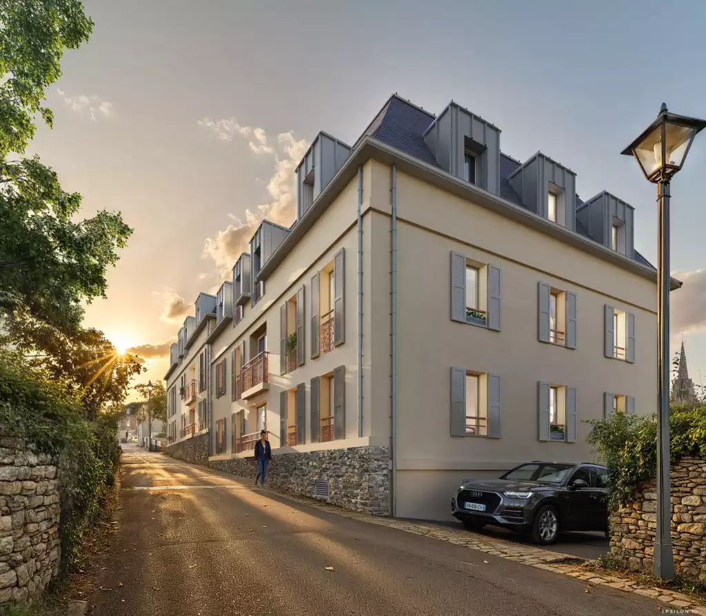 Cette perspective architecturale met en scène la résidence depuis une ruelle en pente, magnifiée par les éclats dorés d'un coucher de soleil. Le rendu souligne le caractère authentique du projet grâce à l'intégration d'un soubassement en pierres sèches qui fait écho aux murets environnants, tandis qu'un lampadaire traditionnel et une silhouette de clocher au loin ancrent le bâtiment dans son histoire locale.