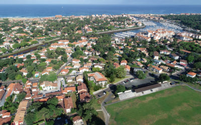 Cette image 3D est une vue aérienne d'une ville côtière ensoleillée. Au premier plan, on peut voir des zones résidentielles avec de nombreuses maisons aux toits de tuiles oranges, entourées d'une végétation dense. Au centre de l'image, un port de plaisance est visible, rempli de bateaux. Il est relié à l'océan par un canal. L'océan, de couleur bleue et calme, s'étend à l'horizon. Une plage de sable s'étend le long de la côte, et on peut voir des bâtiments côtiers derrière elle.
